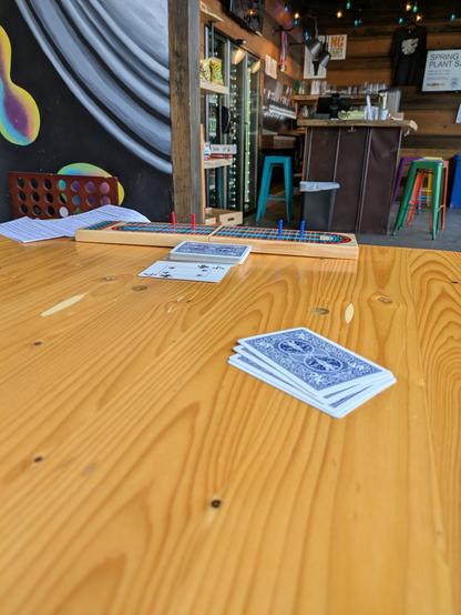 A cribbage board and cards one a wood table in the foreground.  In the background are colorful bar stools, a commercial beer fridge, and bar.