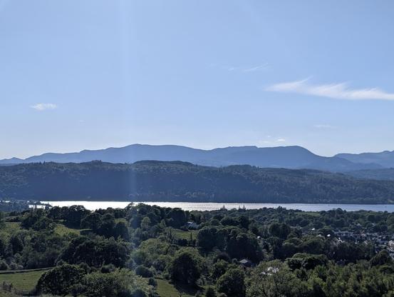 Foreground of fields and trees. Background hills against a blue sky. In the middle, Lake Windermere lit by bright sunshine