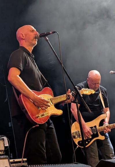 Singer playing guitar next to bassist on stage with a black,smoky background