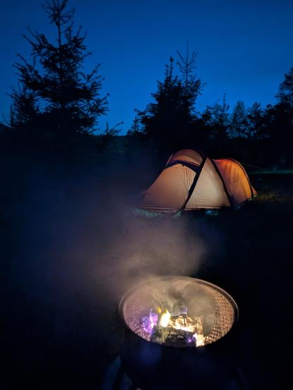 Night time. In the foreground is a fire pit with low flames. Beyond is an orange tent, lit from inside. Black trees behind