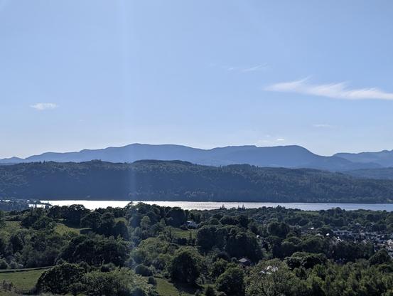 Foreground of fields and trees. Background hills against a blue sky. In the middle, Lake Windermere lit by bright sunshine