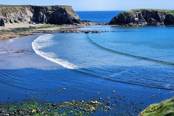 A high-angle shot shows a curved sandy beach nestled between rocky cliffs and the clear blue sea in Annestown, Ireland. Gentle waves roll onto the shore, and seaweed-covered rocks are visible in the foreground.