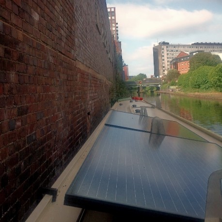 A very long, vertical, red brick wall fills the left side of the photo. The wall goes straight down into the water of the canal – no ledge, no steps, no towpath, nothing to stand or walk on.

The boat's midline is looped through the ring (out of view below the boat's roofline) and then tied to the boat.

On the far side of this rather broad canal is the towpath. This urban canal continues under a bridge in the distance. There are various industrial-age ruins and some newer, residential buildings alongside the canal. This canal is part of Birmingham Canal Network, or BCN. There are some green deciduous trees and bushes on the towpath side.