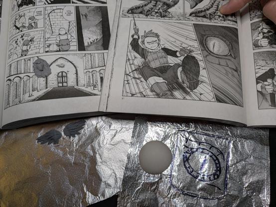A set of polymer clay wings and a giant polymer clay disc sit on aluminum foil. Next to the disc is a rough black marker sketch of a reptile eye, sized to fit an arch outline. 
Above both are 2 manga books opened to pages showing a tiny winged lion wall detail and a close-up of the red dragon's eye, with bonus Chilchuck action shot