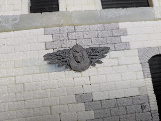 The clay wings with a 3D printed flat lion head glued on, sitting on an unpainted scale model brick wall