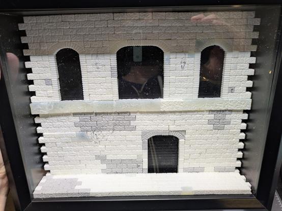 An unpainted scale model brick wall with a small brick floor, sitting inside a diorama box with a plastic "glass" pane