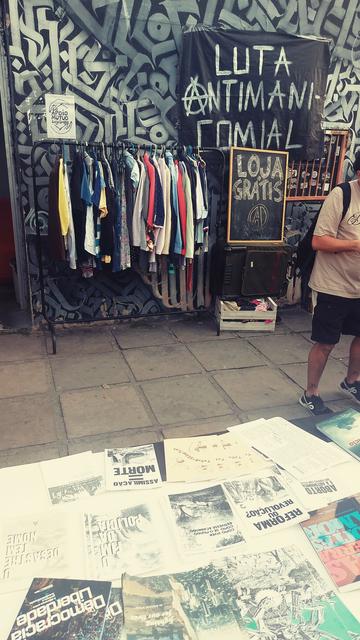 Fotografia de uma banca com diversos zines e livros em primeiro plano. Ao fundo, uma arara de roupas ao lado de um quadro negro onde está escrito: "Loja Gratis". Acima colada numa parede está uma faixa preta escrito: b Luta antimanicomial"