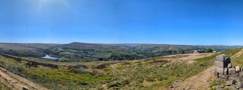 View from a high moorland path across a valley to moorland hills beyond.  In the valley are a reservoir, farms,   golf club, cricket ground, trees, and a sprawling village