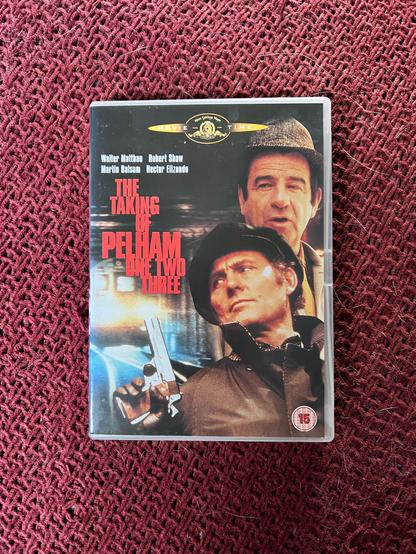 A photo of a DVD copy of the original 1974 movie The Taking Of Pelham One Two Three. The cover has a photo of Robert Shaw at the front, looking to the left and holding a pistol in his right hand. Behind him is a photo of Walter Matthau also looking to the left. The movie title is in red on the left with a photo of a subway train just below it.