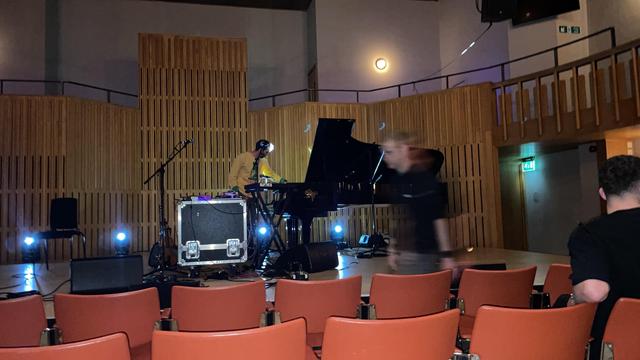 An empty concert hall is shown, featuring a musician at a keyboard next to a grand piano. Stage lights illuminate the space. Some audience chairs are visible in the foreground, with a few people in motion.