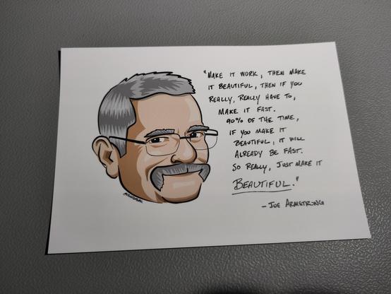 Photo of a postcard with a drawing of Joe Armstrong's face, wearing glasses, a moustache and a smile. The text on the side, probably a quote: "Make it work, then make it beautiful, then if you really really have to, make it fast. 90% of the time, if you make it beautiful, it will already be fast. So really, just make it beautiful"