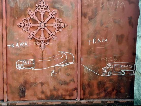 A rust-colored metal gate in Antananarivo, Madagascar, featuring white chalk drawings of two minibuses, commonly used as public transport known as "taxi-be." The minibuses are labeled "TRARA" with stylized motion lines and a hand giving a thumbs-up between them.