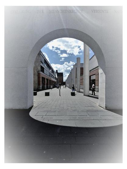 Das Bild zeigt eine architektonische Szene, die durch einen großen, weißen Bogen betrachtet wird. Der Bogen ist Teil eines modernen Gebäudes und bildet einen natürlichen Rahmen, der auf eine breite, gepflasterte Straße hinausführt. Im Hintergrund sind mehrere Gebäude zu sehen, die eine Mischung aus modernen und traditionellen Stilen aufweisen. Die Fassaden sind in verschiedenen Farben gehalten, darunter Weiß und Braun, und einige haben große Fenster.

Die Straße ist von Menschen belebt, die entlanggehen, was auf eine lebendige Umgebung hinweist. Der Himmel ist blau mit einigen weißen Wolken, was auf einen sonnigen Tag schließen lässt. Auf dem Bogen sind die Worte "ERKLARUNG DER MENSCHENRECHTE VEREINTE NATIONEN" zu lesen, was auf eine Verbindung zur Vereinten Nationen und möglicherweise zu einem Ort hinweist, der mit Menschenrechten in Verbindung steht.

Die gesamte Szene ist in einem monochromen Stil gehalten, was die architektonischen Elemente und die Struktur der Umgebung hervorhebt. Die Verwendung von Schatten und Licht verleiht dem Bild Tiefe und Dimension.