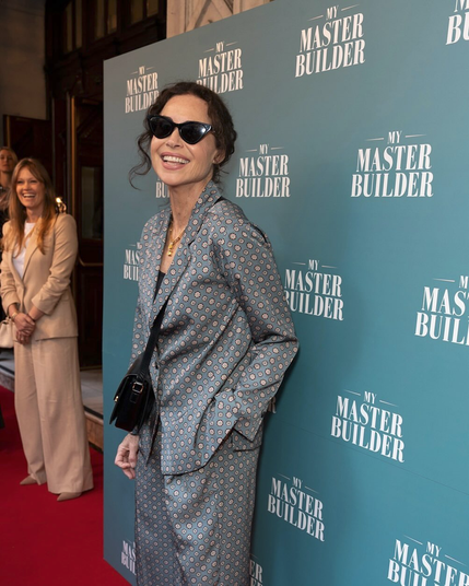 Another photo of Minnie on the red carpet at the opening night party, this time seen from the side. She has one hand tucked into her trouser pocket, and her head is turned to the side to look at the camera with a wide smile. Minnie's friend from the first image stands in the background, looking at something off-camera and laughing.