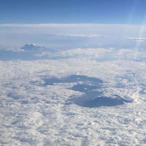 A photo from an airline passenger seat where Mount St. Helens is in the foreground, mostly in shadow, with some ridges poking out by surrounding stratus clouds. Mount Rainier is in the background but only partially covered by clouds. There's some snow on Mount Rainier and the saddle between Point Success and Mt. Rainier peak is easy to see.