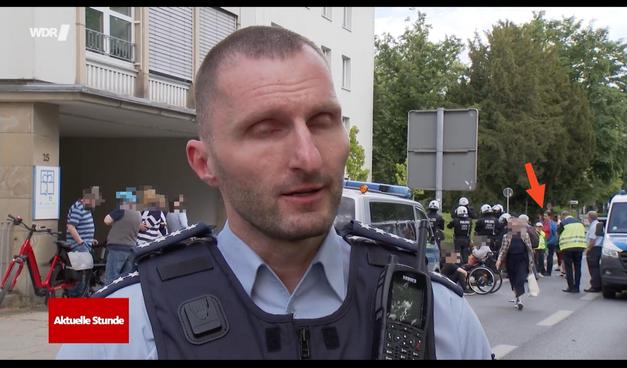 Screenshot aus der Sendung Aktuelle Stunde. Im Vordergrund ist ein Polizist mit Schutzveste, der grade Interview wird. Im hintergrund sieht man Links ein Gebäude, welches der Diakonie sieht, davor sind 4 Personen in einer Gruppe. Rechts hinter dem Polizisten sieht man 2 Polizeiautos. Zwischen den Polizeiautos sind einige Menschen. Etwa 7 Polizisten in voller Einsaztkleidung mit Helmen bilden einen Kreis und setzen so einige Menschen fest. vor dem Kessel sitzen 2 Leute auf dem Boden und eine im Rollstuhl Eine Person geht mit Einkauftaschen richtung Kamera. Rechts von dem Kessel, sind 2 Menschen in Warnwesten und eine Person die Blau/Rot gekleidet ist. Auf die Person in Blau/Roter Kleidung ist ein Pfeil.