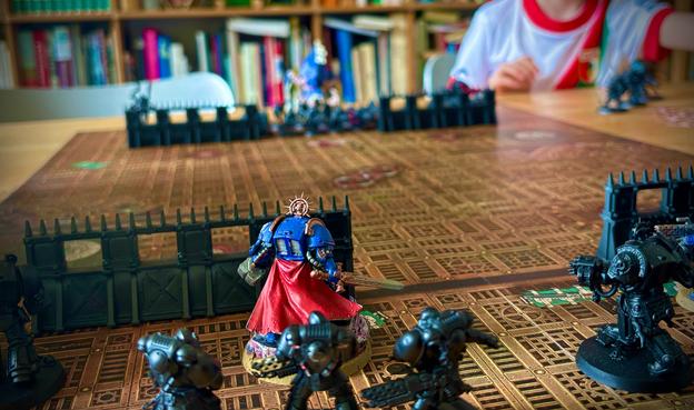 A tabletop wargaming scene showing detailed miniature figures on a game board. The focus is on a vividly painted blue Space Marine with a red cape standing behind a plastic barricade, facing several black armored figures across the board. In the background, a child in a red and white shirt is visible, seemingly orchestrating the game from their side. The setting appears to be a cozy room with bookshelves, giving it a casual but immersive gaming atmosphere.