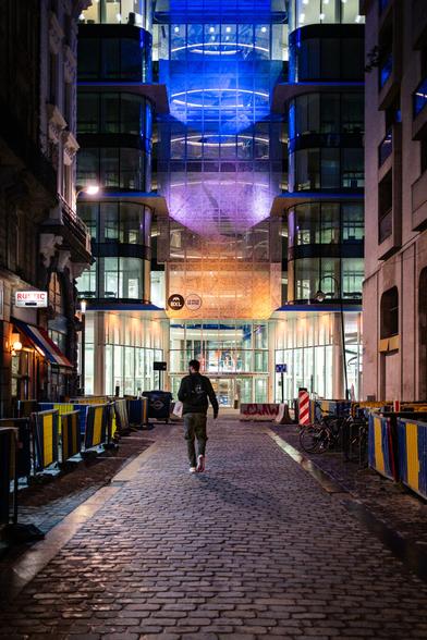 A man seen from the back at night is going down a dark, narrow and empty small street in Brussels towards a tall modern building illuminated with big neon circles of different colors on each floor, creating a science fiction atmosphere.