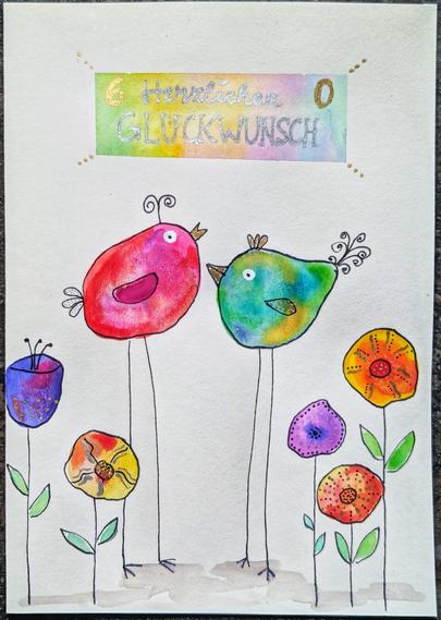 A handmade greeting card is shown on a textured white background. At the top, a rectangular banner with a shimmering silver and gold wash has the handwritten text "Herzlichen GLÜCKWUNSCH" and the numbers "6" and "0". Above and below this banner are small clusters of gold dots.
Below the text, two whimsical birds face each other. The bird on the left is predominantly pink and red with a darker pink wing and a black outlined eye. The bird on the right is a mix of green, blue, and yellow, also with a black outlined eye. Both birds have thin, elongated black legs and small black curled details on their heads and tails.
At the bottom of the card are several stylised flowers in various vibrant colours including blue, orange, yellow, and purple, each with simple green leaves and stems. The overall style is colourful and illustrative.