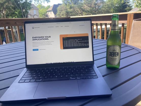 A black MacBook Pro on a black metal garden table flaked by a green bottle of Jever Pilsener. A deck railing is in the background. The screen shows the Apptainer homepage.