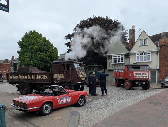 A photo showing an old Triumph Spitfire sports car, and two historic steam lorries on a cloudy morning in an old English town.