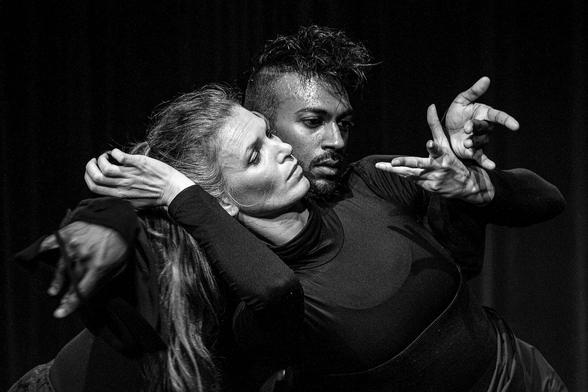 A black and white photo. Two dancers, a woman and a man. The woman bends backwards from right to left, the man the other way around, behind her and close to her. She rests her head on his right shoulder. You can see the dancer down to just below her waist. They are both dressed in black. All you can see of the dancer is his head and hands. His head rests on the woman's left shoulder. Their faces are close together. The woman has white skin and long blond hair in a ponytail. The man is dark-skinned, with a moustache and shaved hair. Their left hands are against each other, their fingers spread. The woman's right arm is bent, her hand along her head. The image is intense, the faces concentrated, sweaty.