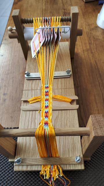 Folding tablet loom made from oak with a tablet woven band in progress. Band is woven from crochet cotton in variegated gold on both sides with centre stripes of lime green, pale blue,deep pink and hot orange