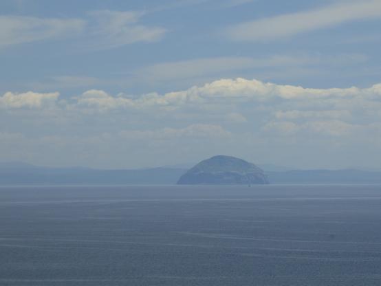 A distant dome-shaped island in a calm sea with a band of grey and white cloud above it and the dark shape of a mountainous landscape far behind it.