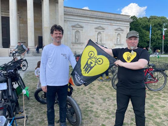 Zwei Männer und die CM Flagge auf dem Königsplatz vor der Glyptothek in München. Einer hat ein CM T-Shirt an (schwarz, gelbes Logo), der andere ein weißes mit dem Logo von Munich Ways