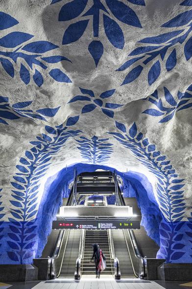 Vertical wide angle view of an ascending escalator group installed in a painted cave, with a white ceiling decorated with blue laurel wreaths patterns, at T-Centralen metro station in Stockholm, Sweden. A lady is about to ascend the middle stairs in the foreground, in the lower part of the image.