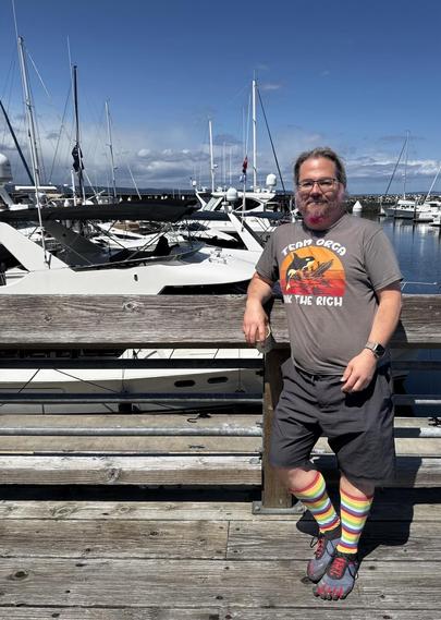 Guy with a pink beard standing in front of a row of yachts at Port of Edmonds. He’s wearing rainbow socks and a shirt saying “Team Orca - sink the rich” showing an orca swamping a super yacht. He has a shit eating grin on his face.