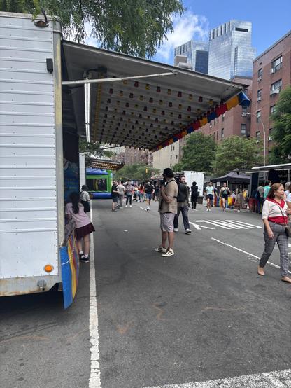 Side view of an amusement game at the 9th Ave Food Festival and street fair. The light-up overhead awning from the game is seen, with rows of dark bulbs. Across the street, apartment buildings, in the distance, high rises and sky.