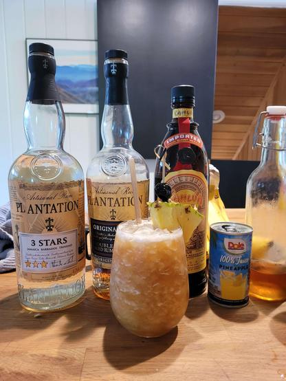 A juicy cocktail, served in a glass full of crushed ice and garnished with a pineapple wedge and a cherry, set in front of two bottles of rum, a bottle of Kahlúa, a can of pineapple juice and a bottle of cinnamon syrup.