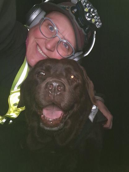 Night time selfie of a person and a chocolate brown Labrador with greying snout. The person is wearing a black jacket, yellow safety harness, red cap, silver headphones and blue-rimmed glasses. On their cap rests a headstrap torch in off position. Their free hand is around the dog's shoulders. Both are facing the camera. The dog's mouth is slightly open, showing off some mlem. The person's head is resting on the dog's head. They're smiling, grateful for their time spent together.