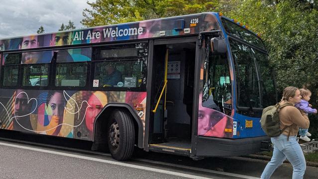 A colorful city bus with the words "All Are Welcome" above the windows and a kaleidoscope of multihued people below the windows.