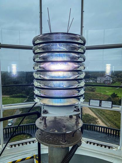 A close-up view of a modern lighthouse beacon, captured from inside the lantern room at the top of the tower. The beacon is a VLB-44 LED optic composed of stacked, circular lenses, each ring shimmering with reflections from the white light glowing at the center. Thin metal rods extend upward from the top of the fixture. The base of the beacon is mounted on a sturdy metal platform secured with bolts. Through the large glass windows surrounding the beacon, a green landscape is visible under a cloudy sky.