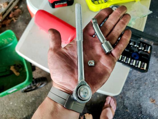 A broken ratchet wrench and bench extension piece in the photographer's dirty left hand. A socket set, Bluetooth speaker, and the photographer's right foot are in the background.