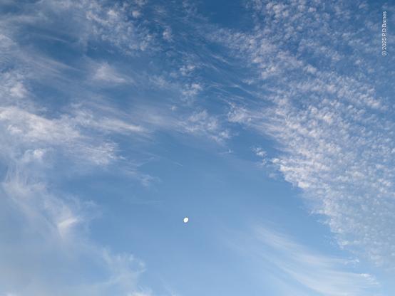 A gibbous moon sits in a relatively clear patch of blue sky, surrounded by lines of white stratus cloud that have been blown into wisps to the left, while remaining intact as tiny little puffballs on the right.