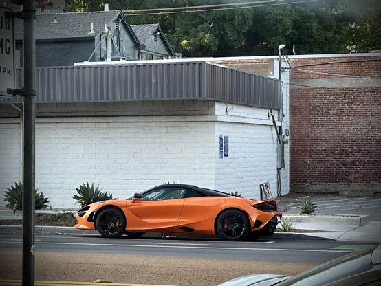 A vibrant orange sports car is parked on the street next to a textured white wall and a brick building. The car displays sleek lines and a low profile, set against a backdrop of greenery and urban architecture.
