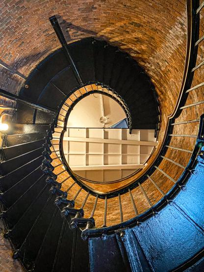 A black metal spiral staircase winds upward inside the round brick interior of a lighthouse. The photo is taken from below, looking straight up through the center of the staircase. The inner brick walls curve around the staircase. The stairs are bordered by thin vertical metal railings, and warm lighting from the left casts a soft glow on the bricks and metal.