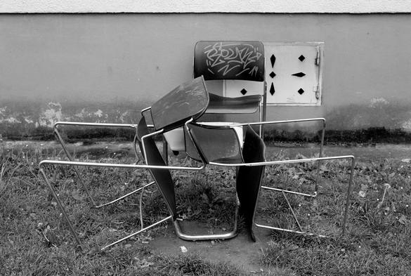 The black and white image shows a pile of stacked and overturned chairs on a patch of grass against a wall. The central chair has graffiti on its backrest, and the others are entangled in a haphazard, sculptural arrangement. A small access panel with diamond-shaped cutouts is visible on the wall behind.