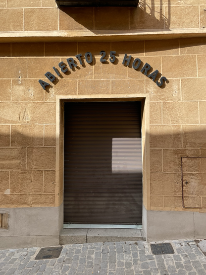 A tan stone wall with a metal door that is shuttered. Over the door, text reads “Abierto 25 horas.”