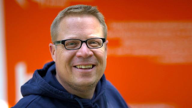 Portrait von Dominic Kirchner-Schmidt. Nahaufnahme eines lächelnden Mannes mittleren Alters mit kurzen, blonden Haaren und einer Brille. Er trägt einen dunkelblauen Kapuzenpullover. Der Hintergrund ist unscharf und zeigt eine orangefarbene Wand mit unleserlichem Text.