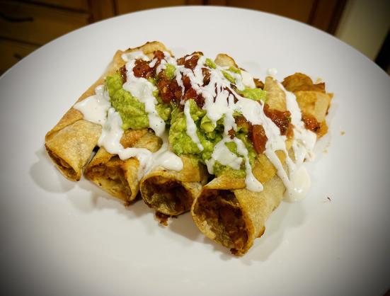 Four taquitos close together on a plate. There is guacamole and salsa piled across the middle of them, with the crema drizzled across diagonally.