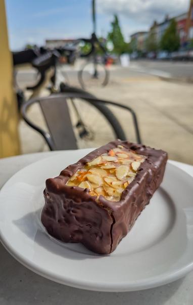 A chocolate-covered dessert with a nutty filling, sitting on a white plate. In the background, a bicycle and urban scenery are visible