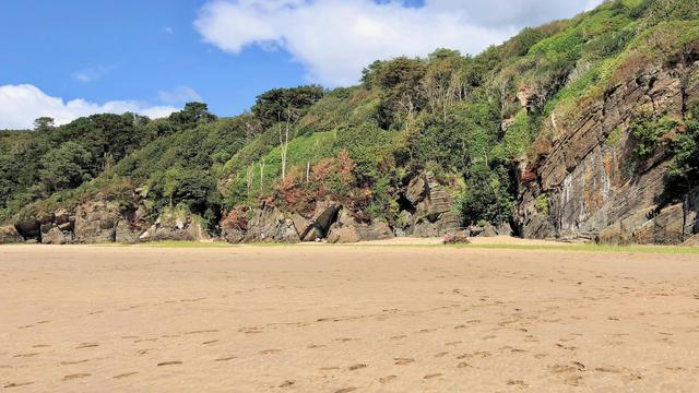 The picture showcases a serene coastal landscape featuring a wide, sandy beach that stretches into the distance. On the left side, there’s a lush, green forested area atop rocky outcrops, adding a natural contrast to the sandy foreground. The beach gently curves to the right, where shallow water reflects part of the sky. In the distance, rolling hills and mountains create a beautiful backdrop, partially covered by dramatic, billowing clouds that dominate the sky. The lighting suggests a partly sunny day, casting soft shadows across the landscape.