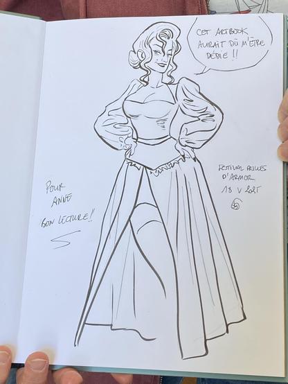 A signin illustration of Lady d’Arcy in a historical dress with oversized sleeves, striking a confident pose. The image includes handwritten text in French expressing that the art book should have been dedicated to her. The context suggests it is part of a festival.