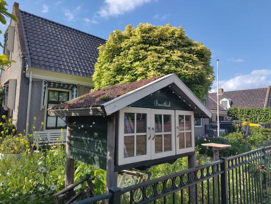 Vrij flinke minibieb voor een huis met kastanjeboom en achter een tuinhek: twee verdiepingen hoog, drie deurtjes, een driehoekig dak waar sedum op groeit.