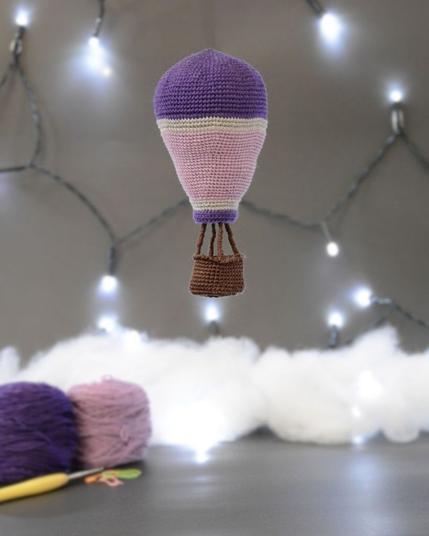 Montgolfière crochetée suspendue dans un décor gris avec des guirlandes lumineuses. Au sol, légèrement en arrière plan, pelotes de laine violette et lilas, crochet et nuages en ouate.