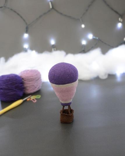 Montgolfière crochetée posée dans un décor gris avec des guirlandes lumineuses. Au sol, légèrement en arrière plan, pelotes de laine violette et lilas, crochet et nuages en ouate.